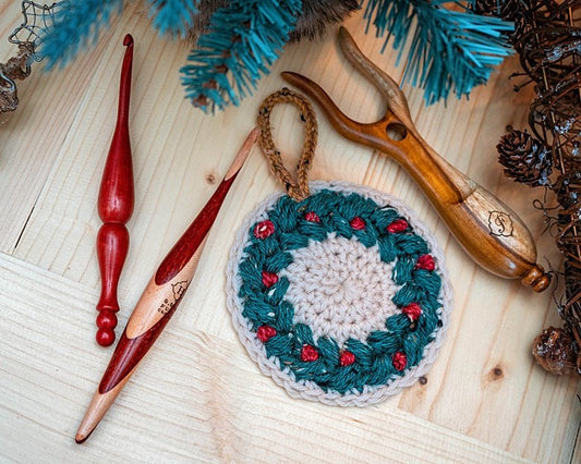Green and cream crochet wreath ornament with red berries on wood beside wooden crochet hooks and pine