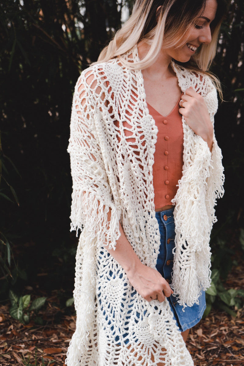 A woman wearing a white shawl decorated with the intricate crochet pineapple stitch, with fringed edges. She stands in an outdoor green space, surrounded by greenery and sunshine.