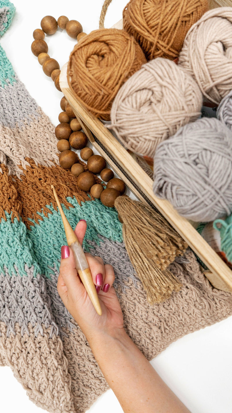 Hand holding a wooden crochet hook over a striped blanket beside a tray of brown, beige and gray yarn