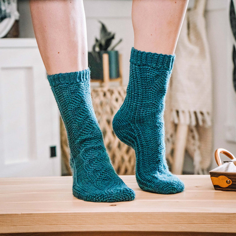 All Cabled Up In Sunshine Socks crochet pattern: teal crochet socks with cabled texture worn by a person standing on tiptoe on a wooden table