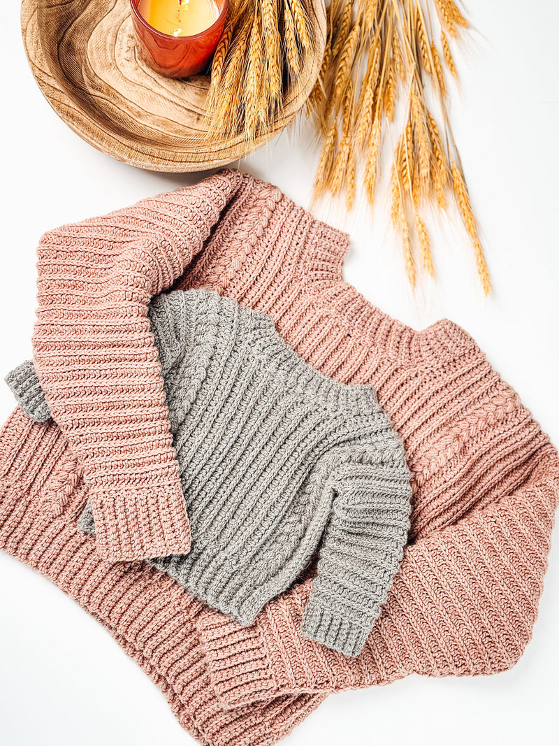 Gray crochet child sweater laid on a larger pink one with wheat stalks and a candle on white