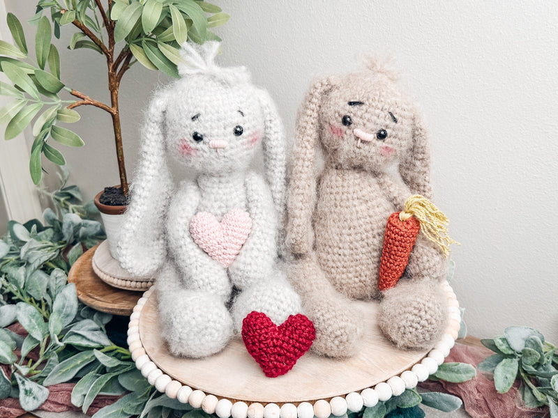 Be My Bunny crochet pattern: two fuzzy crochet bunnies, light gray with a pink heart and tan with a carrot, sitting on a wooden tray with a red heart