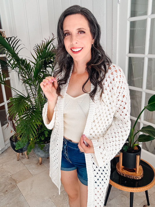 A woman with long dark hair wearing a white crochet cardigan, white top, and blue denim shorts stands indoors in front of a plant and a glass door, smiling at the camera.