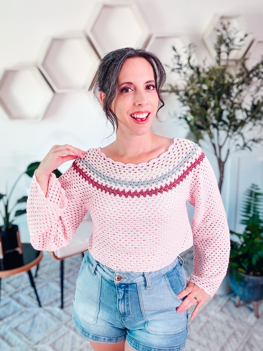 A person wearing a pink crochet top and denim shorts stands indoors smiling. There are hexagonal wall shelves, plants, and a small table in the background, adorned with unique jewelry and vintage clothes.