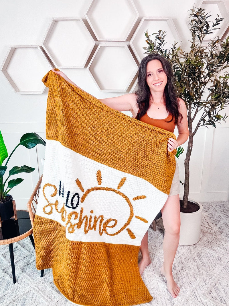 Hello Sunshine Crochet Blanket & Pillow pattern: mustard and white crochet blanket with a sun and script lettering held up by a smiling woman in a bright room