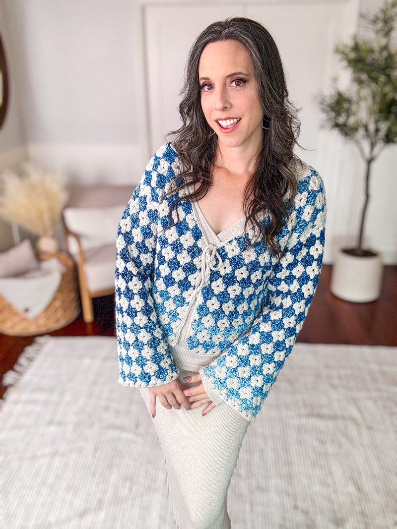 A woman with long dark hair, wearing a blue and white crocheted cardigan—a handmade gift—stands indoors on a light-colored rug with a plant in the background.