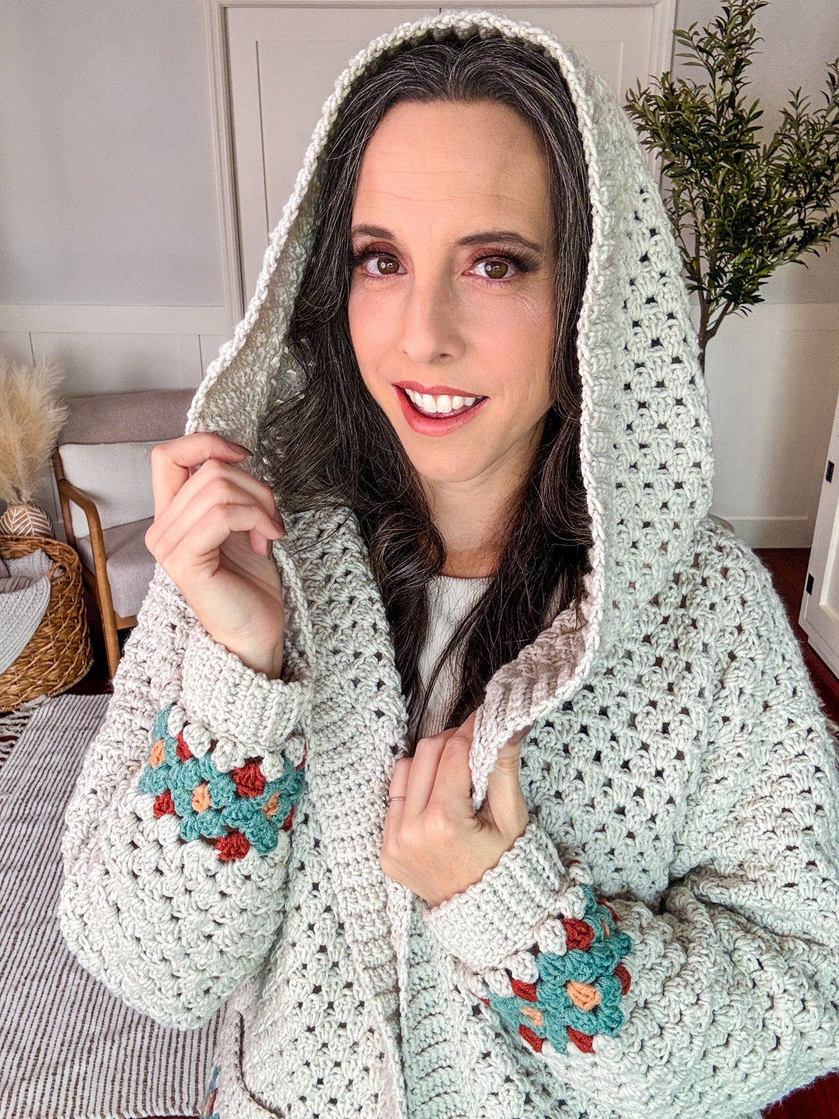 A person with long hair wearing a crocheted sweater with multicolored accents and a hood stands indoors, holding the hood up with one hand and smiling at the camera, showcasing their handmade gift.