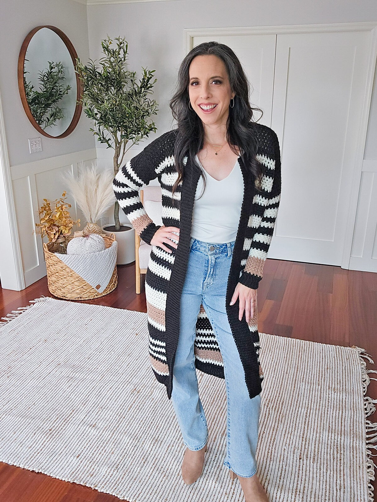 A woman stands in a room with hardwood floors and neutral decor, wearing a black and white striped cardigan, white top, blue jeans, and tan shoes. She is smiling with one hand on her hip, looking as if she just received a handmade gift.