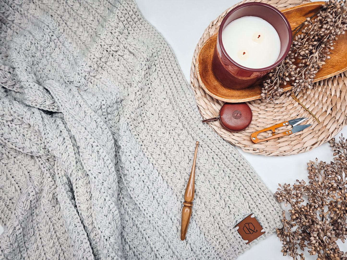 Gray knitted blanket with a wooden crochet hook, a red candle, dried flowers, scissors, and a tape measure on a woven mat—perfect elements for creating the ultimate handmade gift.