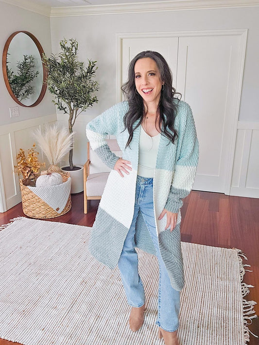 A woman stands in a living room wearing a color-blocked cardigan, jeans, and boots. Adorning her outfit is unique jewelry that catches the light. The room features hardwood floors, a round mirror, and a decorative plant.