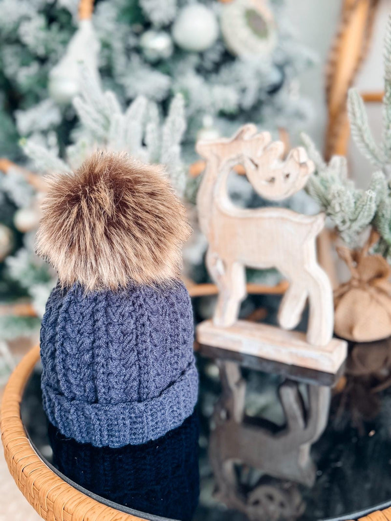 A crocheted blue hat with a faux fur pom-pom, resembling a handmade gift, sits on a glass table next to a wooden reindeer decoration. A decorated Christmas tree glows softly in the blurred background.
