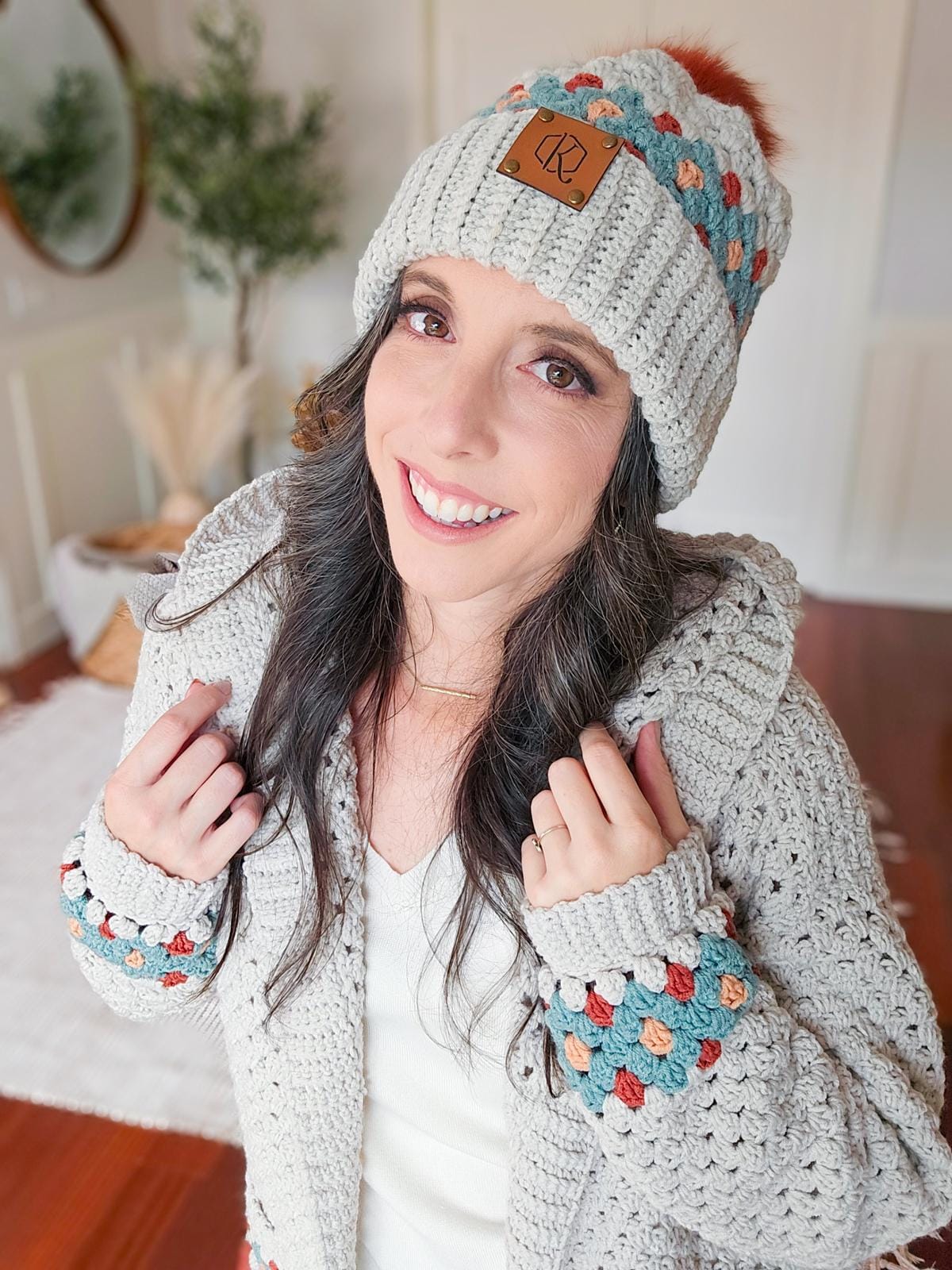 Woman in a light gray crochet sweater and crochet hat with multicolored accents, smiling indoors, showcasing unique jewelry.
