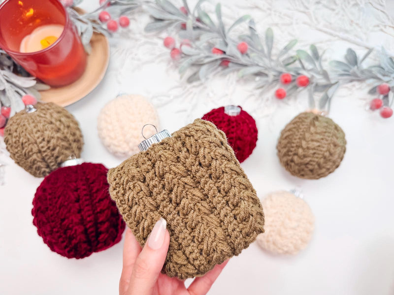 A hand holds a crochet brown ornament, reminiscent of vintage clothes. Other knitted ornaments in red, beige, and brown adorn a white surface alongside a candle and decorative greenery, creating the perfect handmade gift setting.