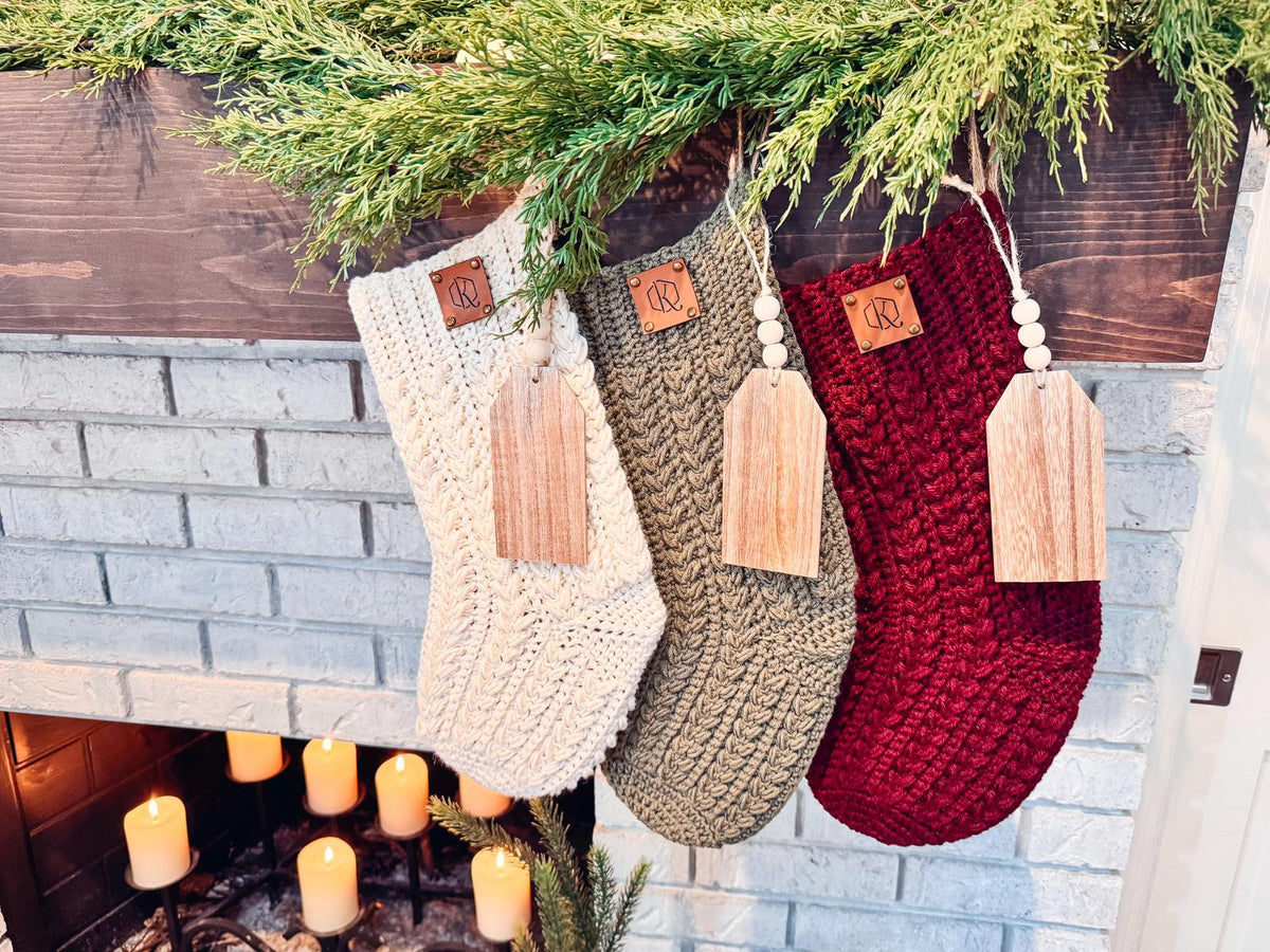 Three crochet stockings in cream, green, and red hang from a wooden mantel adorned with greenery, above a fireplace with lit candles. These handmade gifts perfectly capture the cozy holiday spirit.