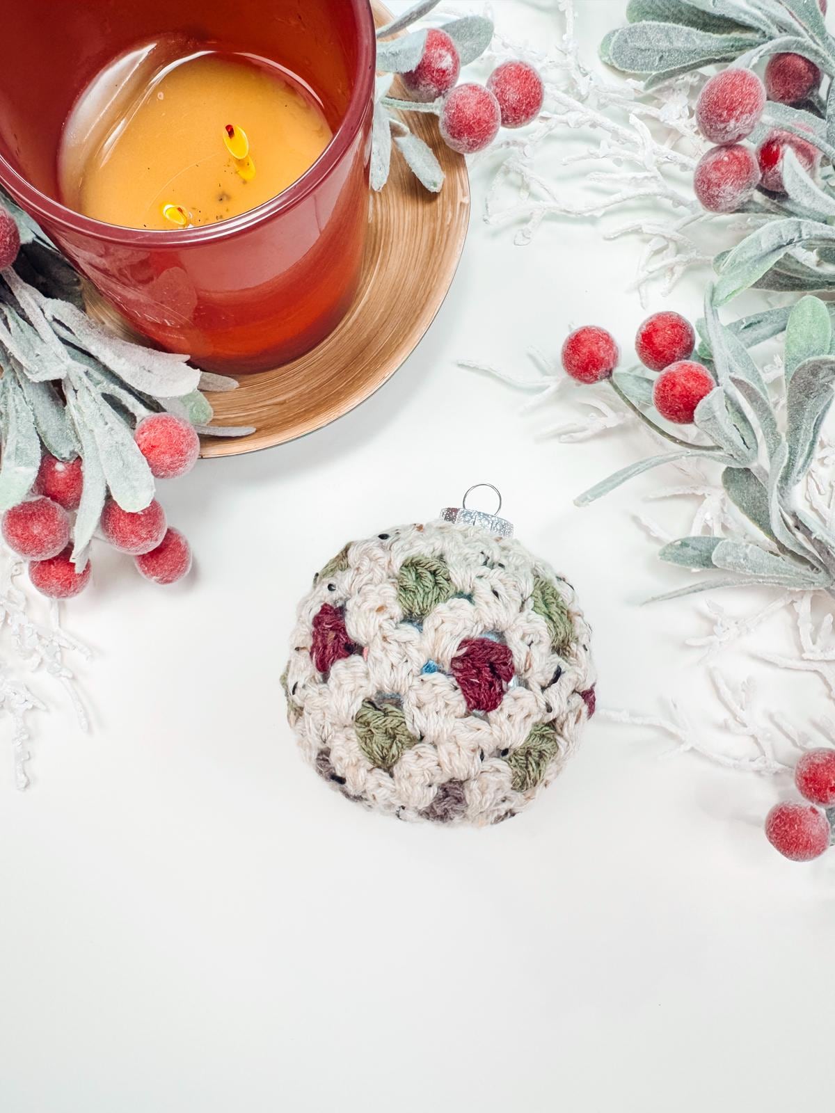 Crocheted ornament with multicolored patterns next to a lit orange candle in a wooden holder, surrounded by frosted greenery and red berries on a white surface—a perfect handmade gift for those who appreciate artisanal charm.