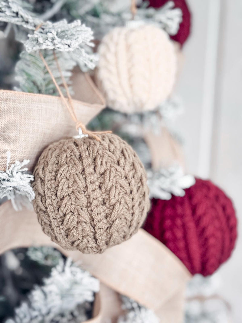 Close-up of a Christmas tree adorned with crocheted ornaments in beige, cream, and red, accented by burlap ribbons and frosted branches, giving it the charm of a handmade gift.