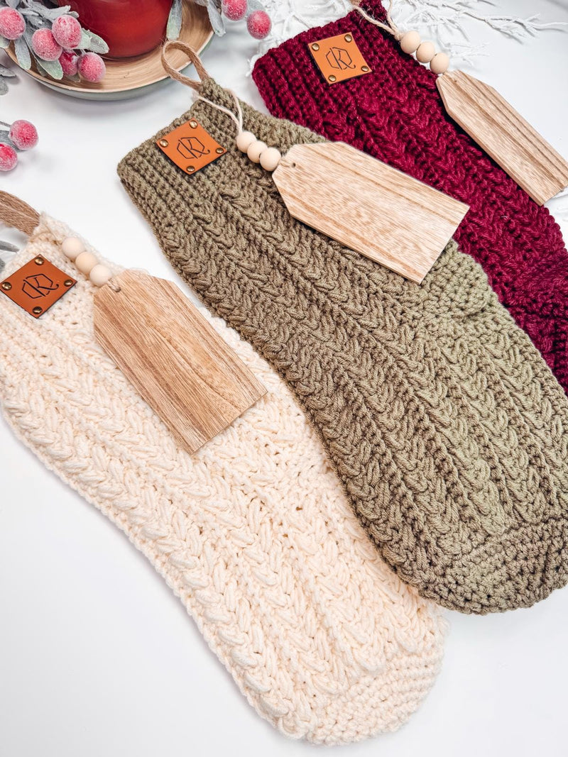 Three crochet stockings in cream, olive green, and burgundy are placed in a row, each adorned with a wooden name tag and leather patch. Perfect as a handmade gift, these stockings add a touch of charm and warmth to any holiday décor.