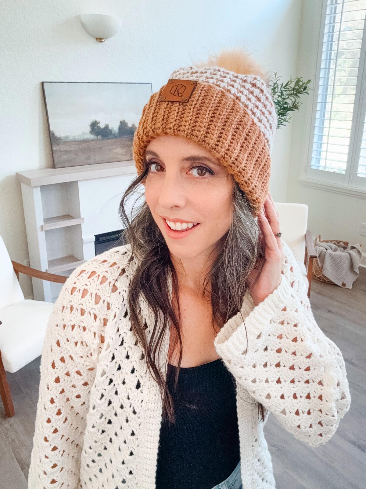 A woman adorned in unique jewellery, wearing a knit hat and crochet sweater, stands indoors with a warm smile. The backdrop features a cozy chair, an elegant wall painting, and a lush plant.