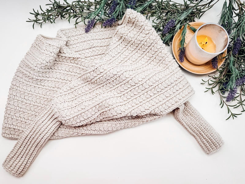 A beige crocheted sweater scarf is folded on a surface, surrounded by sprigs of lavender and a lit candle in a glass holder on a wooden tray.