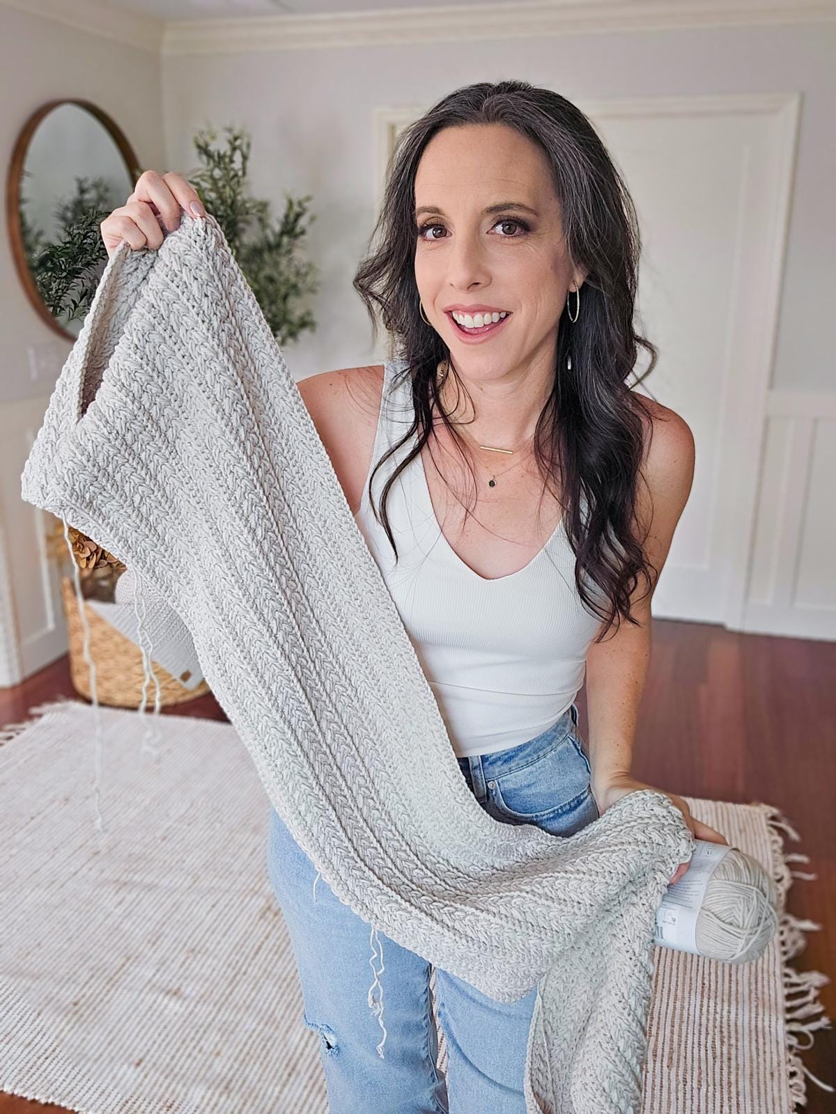 A woman displays a light gray crocheted piece of fabric, standing in a room adorned with a rug and decorative plant, offering it as a charming handmade gift.