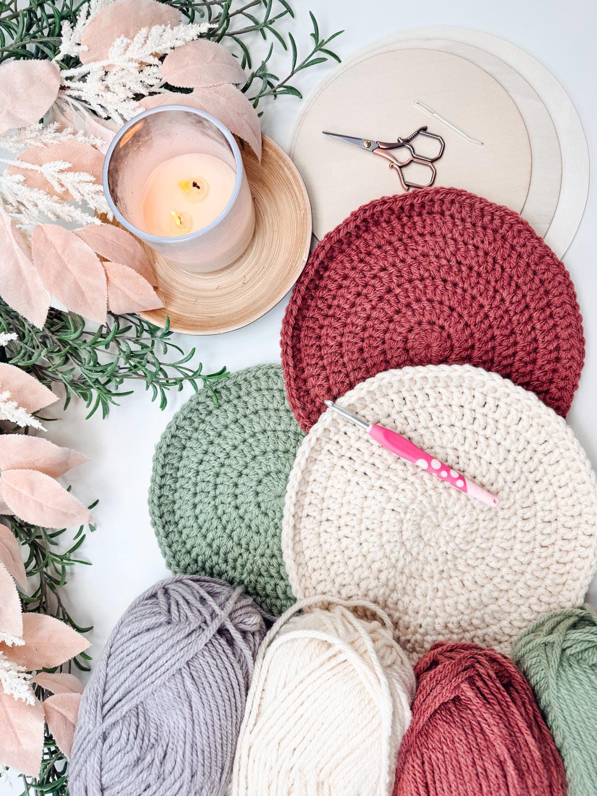 Crocheted placemats in red, green, and beige accompany yarn balls, a crochet hook, scissors, round wooden boards, greenery, and a lit candle on a white surface.
