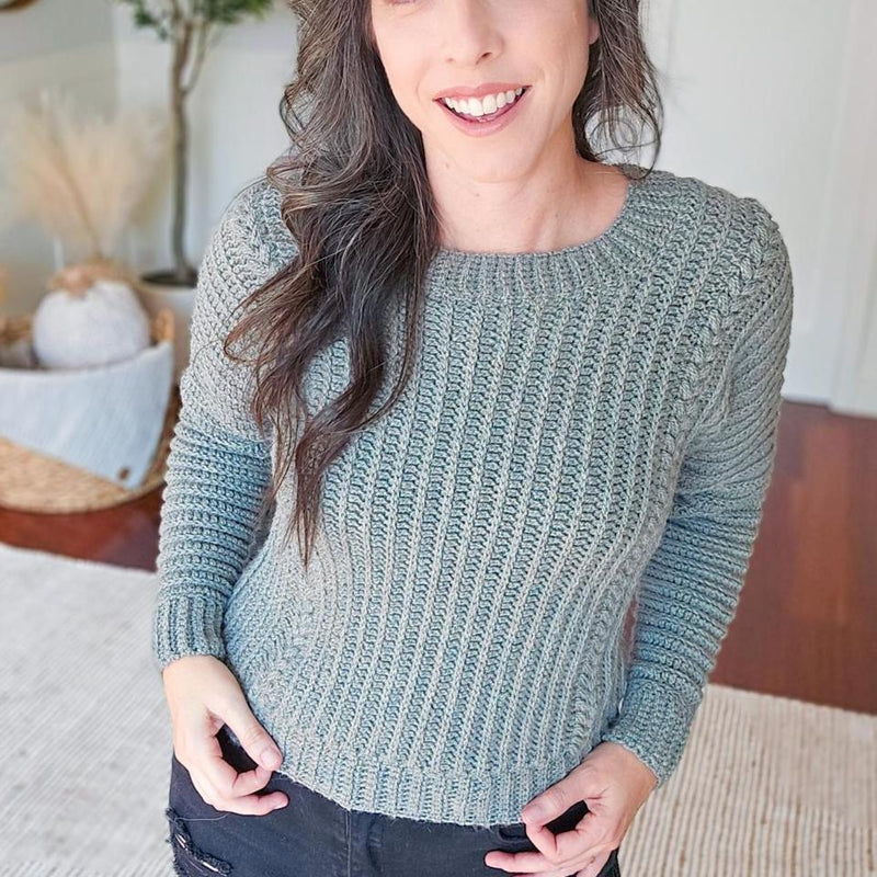 Autumn Wheat Adult Sweater crochet pattern: sage green ribbed crochet sweater with braided sleeve detail worn by a smiling woman with black jeans