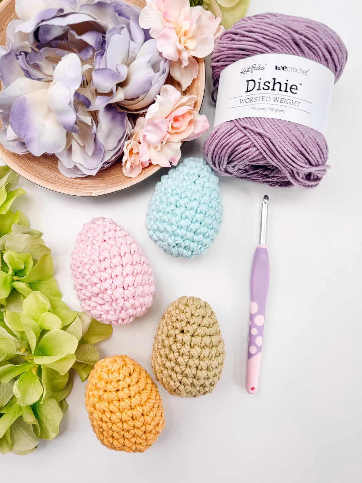 Crochet yarn, a crochet hook, and flower bouquet are arranged with four crochet eggs in pastel colors on a white background, creating the perfect setting for a charming handmade gift.