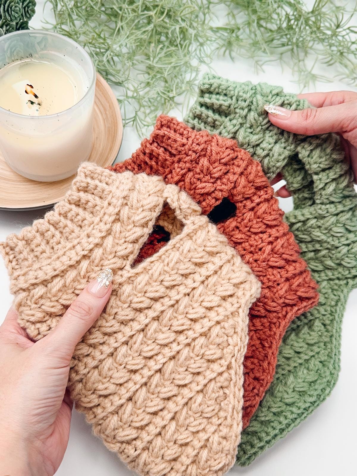 Hands holding three crochet dog sweaters in beige, orange, and green—perfect as a cozy handmade gift; a lit candle and green foliage add warmth in the background.