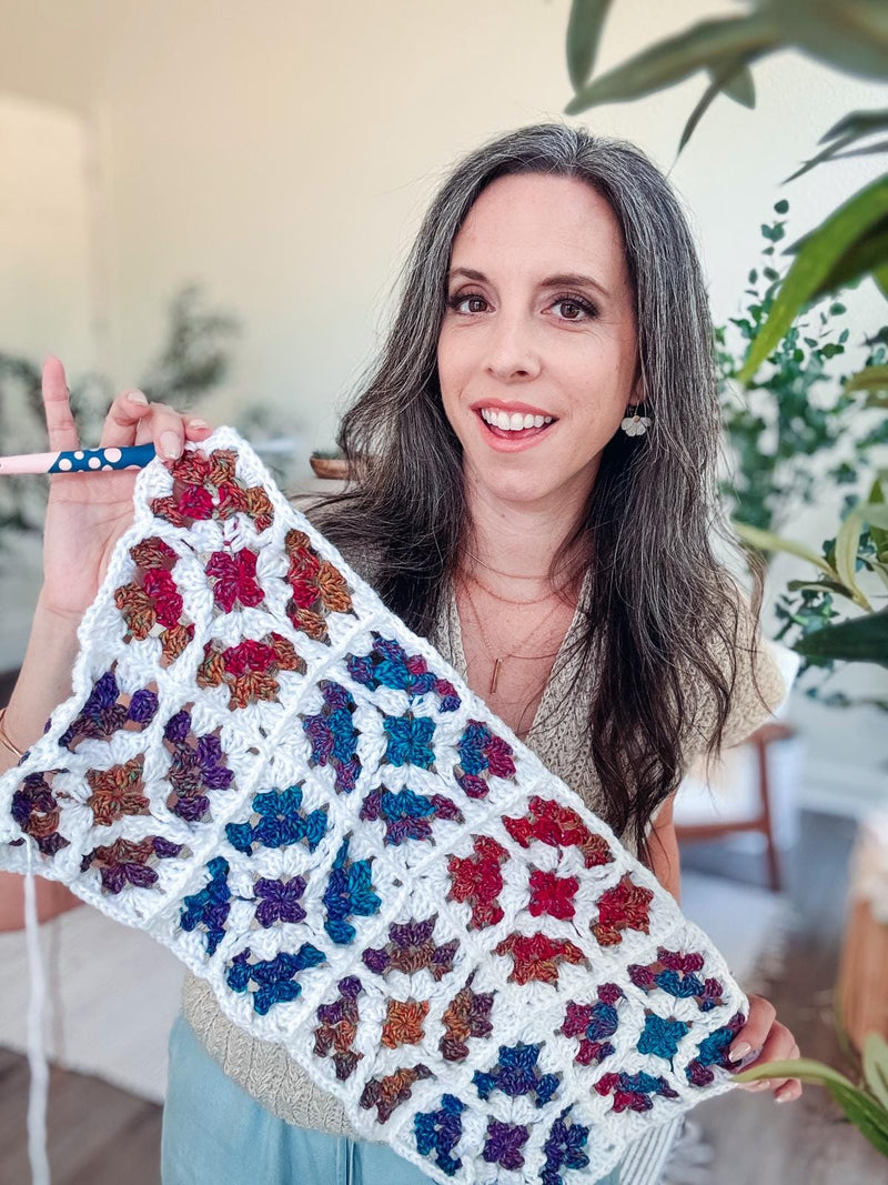 Smiling woman holding a strip of white crochet granny squares with jewel-tone centers and a crochet hook