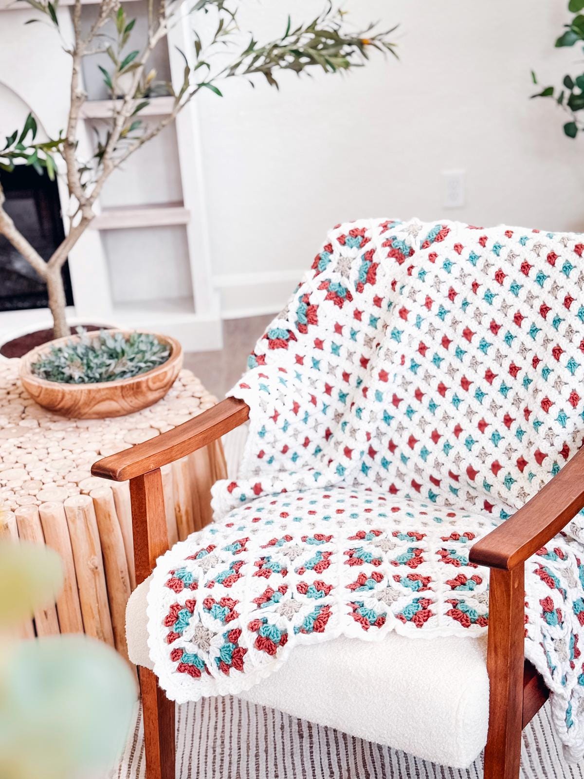 A wooden chair with a crocheted blanket in red, teal, and white draped over it sits beside a round wooden side table with a potted plant—perfect for relaxing or displaying vintage clothes and handmade gifts.