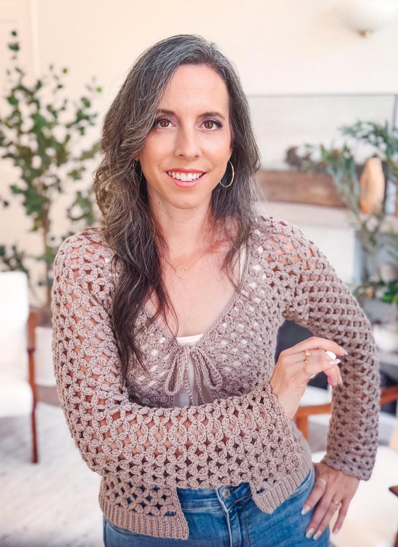 A woman with long brown hair and a crochet sweater smiles at the camera indoors, standing in a bright room with plants, neutral decor, and unique jewellery accenting her style.