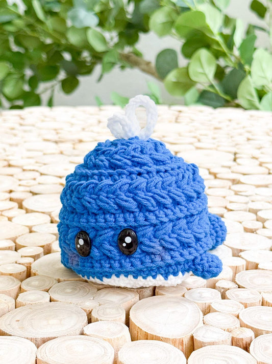 A small, blue crocheted whale with black button eyes sits on a surface of light wooden discs, making a charming handmade gift. Green leafy plants in the background add to its appeal.