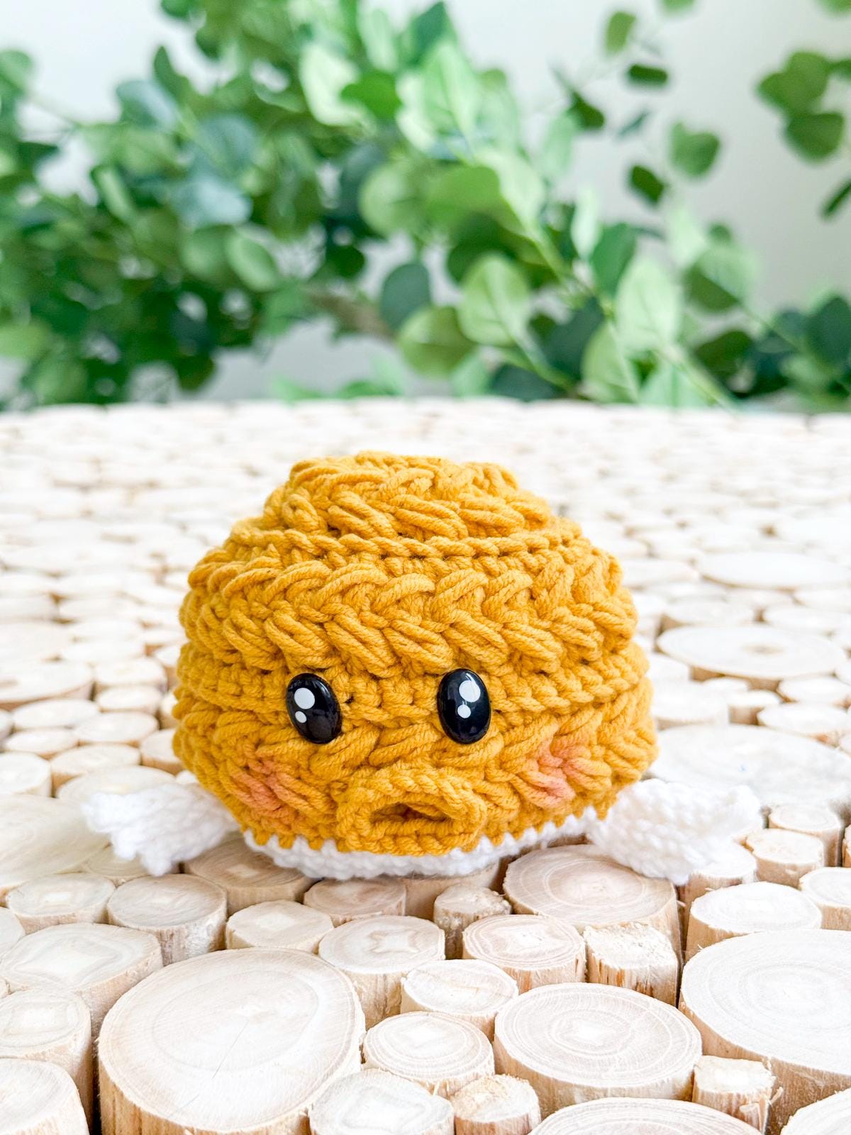 A small, crocheted yellow plush with black eyes and white arms sits on a surface made of cut wooden logs, with green plants in the background—a charming handmade gift sure to delight any collector.