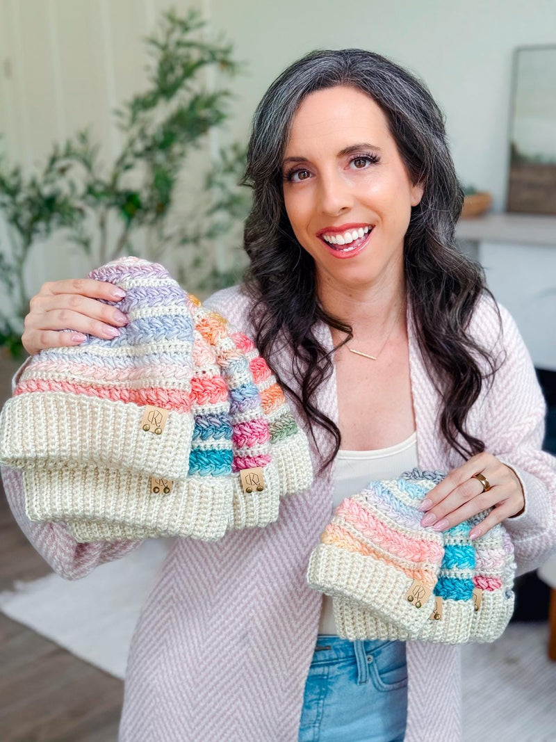 Several pastel striped crochet beanies held by a smiling woman with long dark hair in a pink cardigan