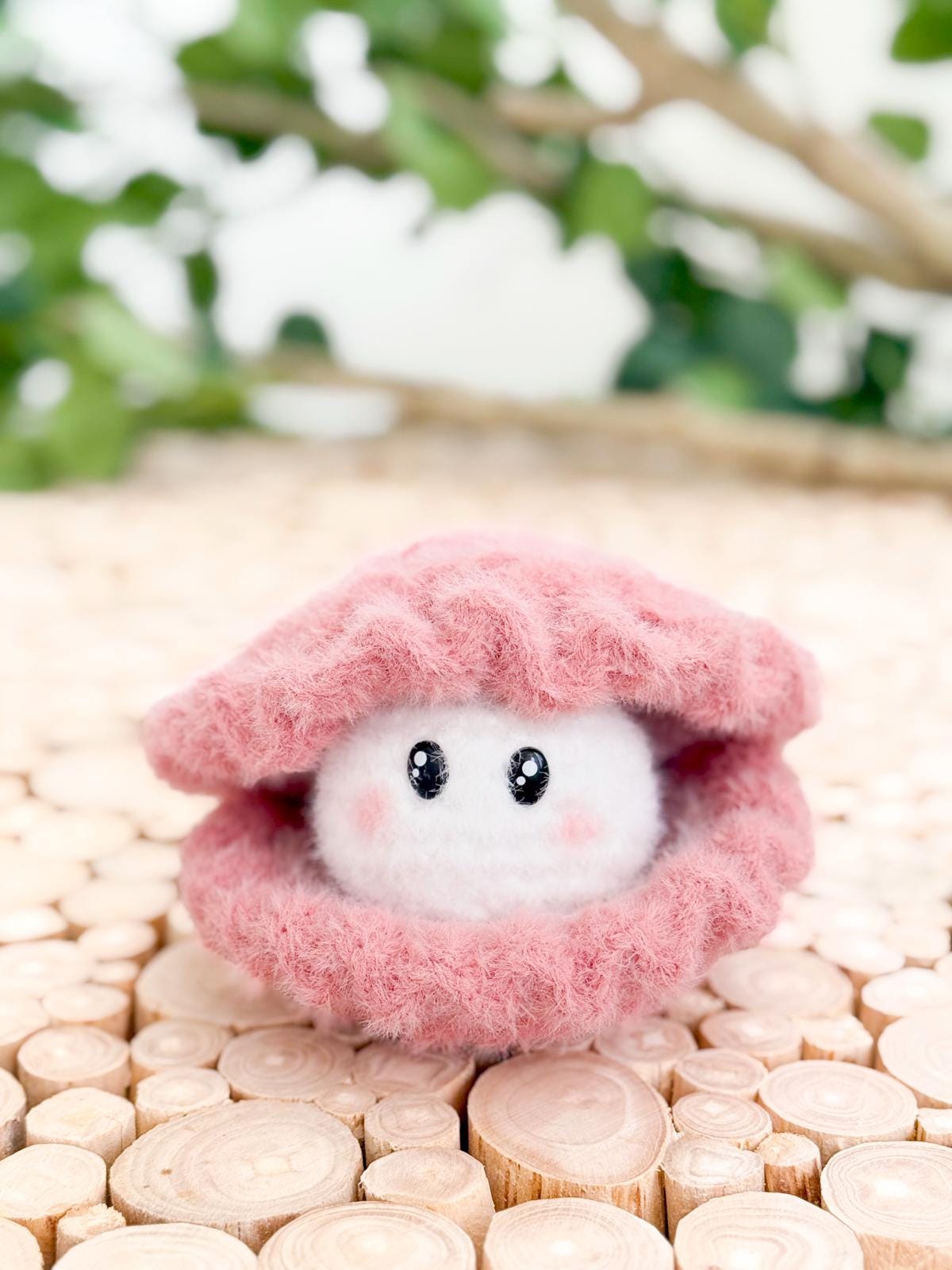 A small plush crochet toy resembling a white pearl with eyes and cheeks sits inside a pink, fuzzy clam shell on a wooden surface, making it an adorable handmade gift. Green leaves add charm to the background.