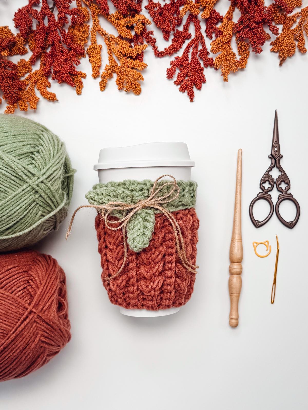 A takeaway coffee cup with a crocheted orange and green cozy sits on a white surface, surrounded by yarn, crochet tools, scissors, and autumn leaf decorations—perfect inspiration for a handmade gift or unique jewellery project.