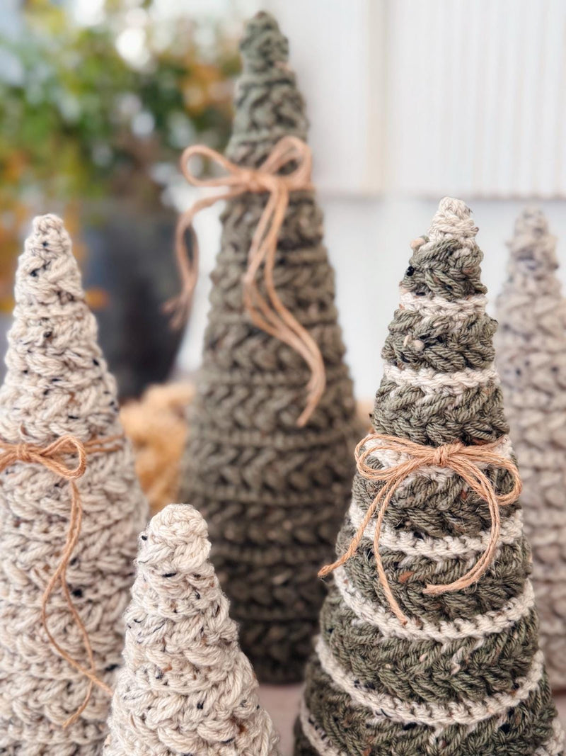 Several handmade crocheted Christmas trees in neutral tones, some decorated with twine bows, are arranged closely together on a surface—a charming handmade gift perfect for adding warmth to your holiday décor.
