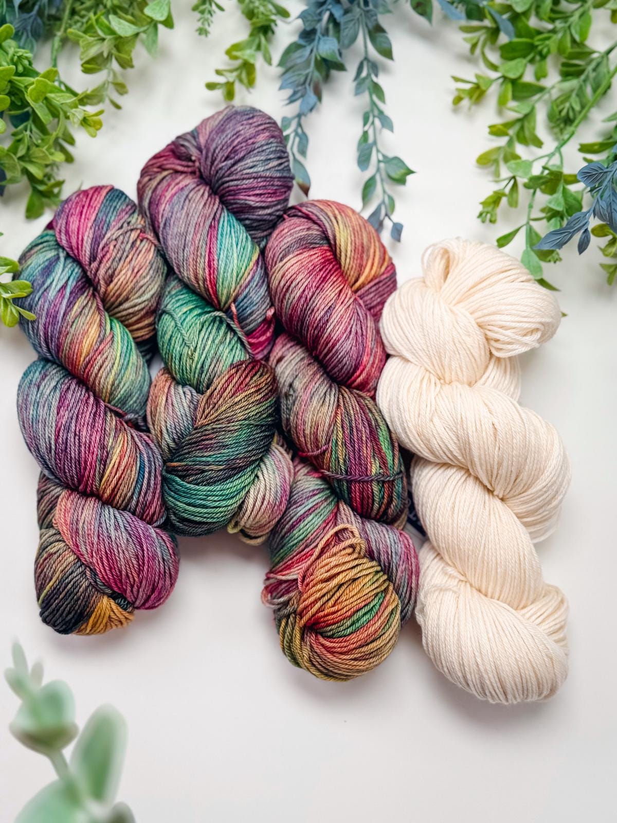 Four skeins of yarn, three multicolored and one cream, are arranged side by side on a white surface with green foliage—perfect for creating a handmade gift or pairing with vintage clothes for a cozy, stylish look.