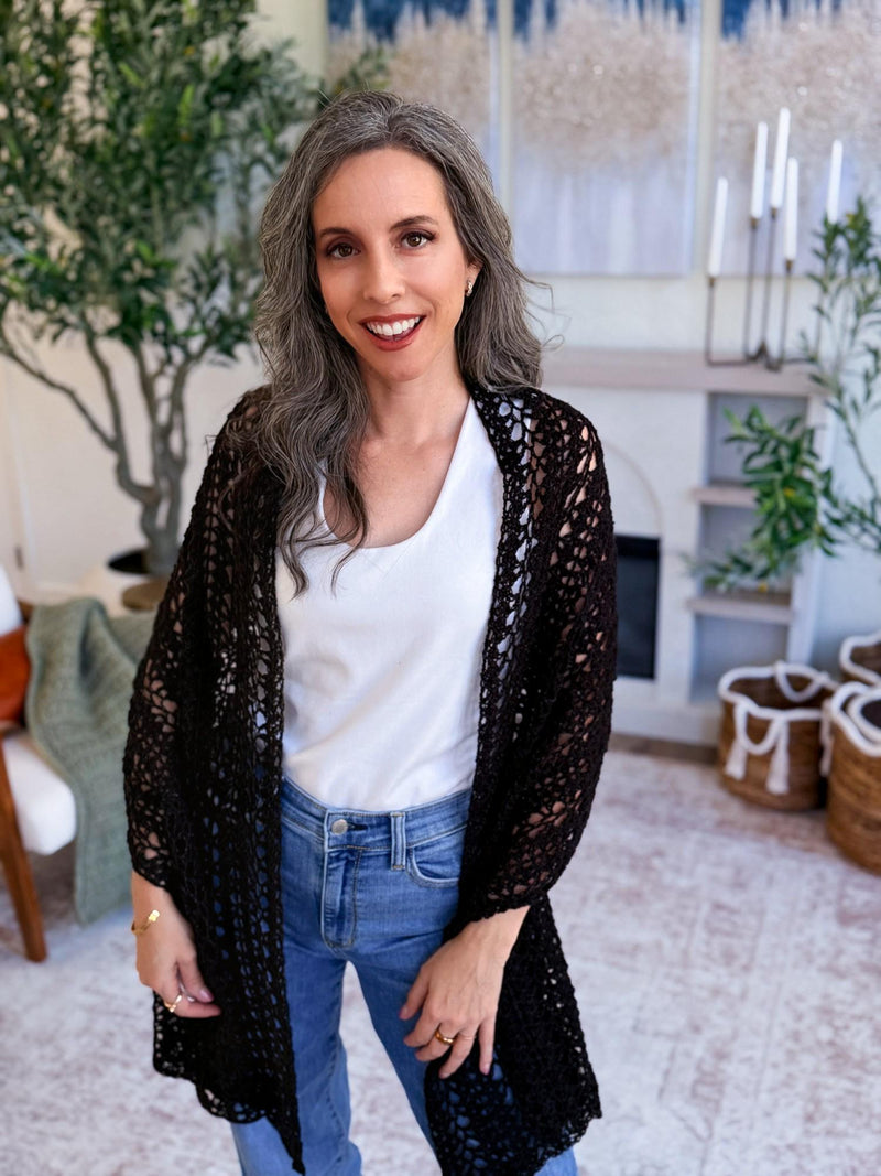 Black crochet mesh wrap worn open like a cardigan over a white top and jeans by a smiling woman indoors