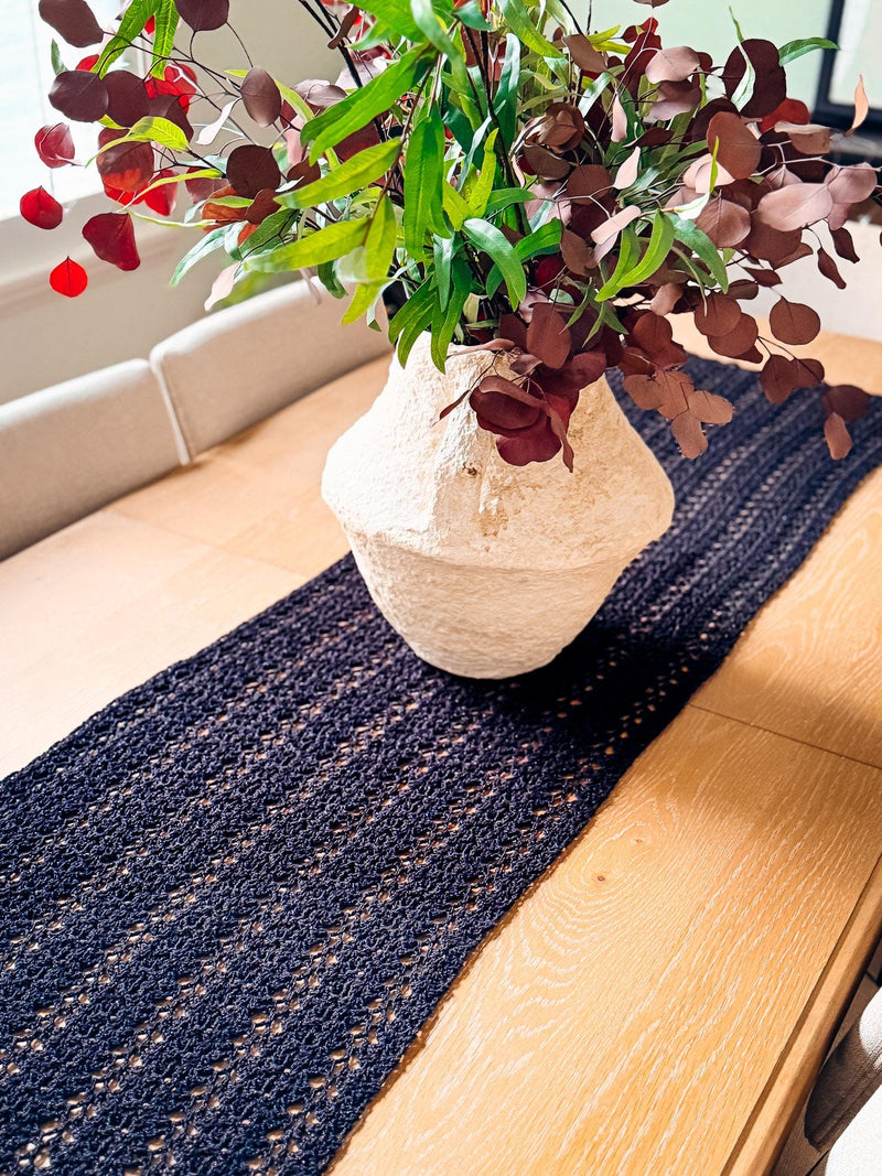 Black lacy crochet table runner on a wooden dining table with a cream vase of green and burgundy leaves