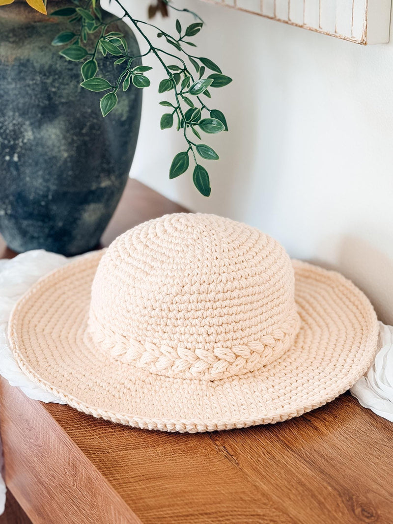 Cream crochet sun hat with braided band resting on a wooden table beside a dark vase and greenery