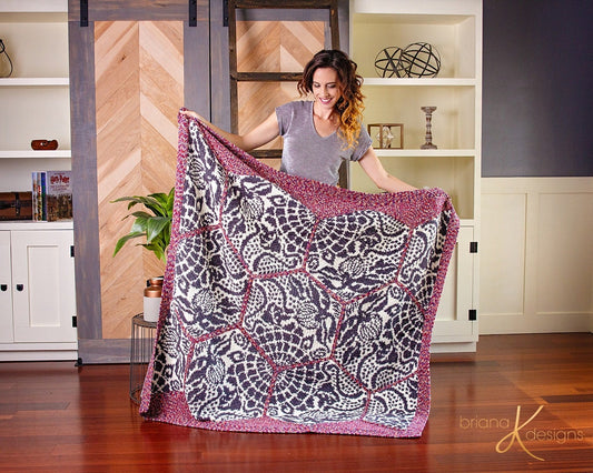 The Gaudi Sidewalk Blanket crochet pattern: purple and cream hexagon crochet blanket with pink trim held open by a woman in a living room