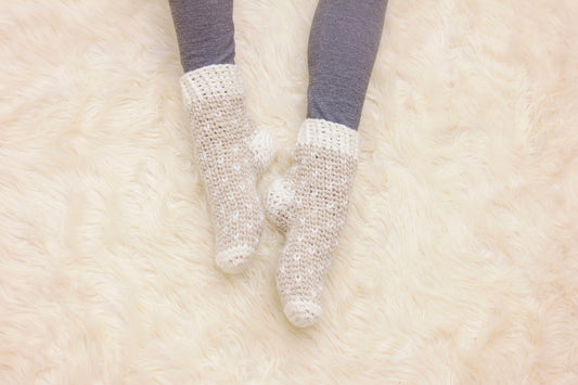 Comfy Cozy Winter Crochet Slipper Sock pattern: oatmeal and cream crochet slipper socks worn with gray leggings, feet resting on a white faux fur rug