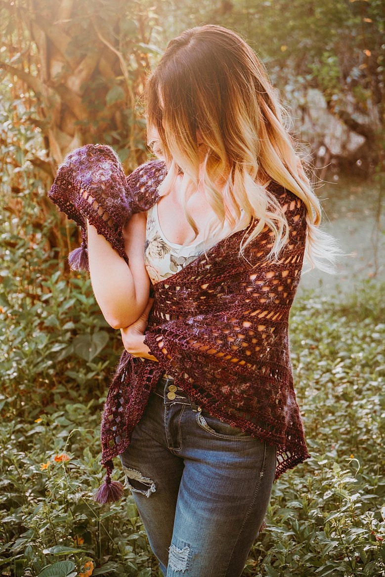 A person stands outdoors, wearing a floral top and jeans, holding a purple crocheted shawl—a charming handmade gift that adds an elegant touch.