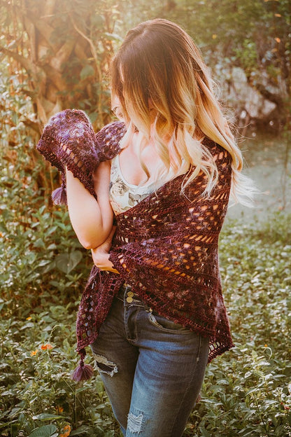 A person stands outdoors, wearing a floral top and jeans, holding a purple crocheted shawl—a charming handmade gift that adds an elegant touch.