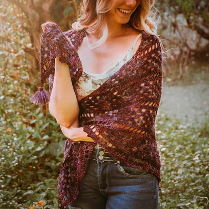 A person stands outdoors, wearing a floral top and jeans, holding a purple crocheted shawl—a charming handmade gift that adds an elegant touch.