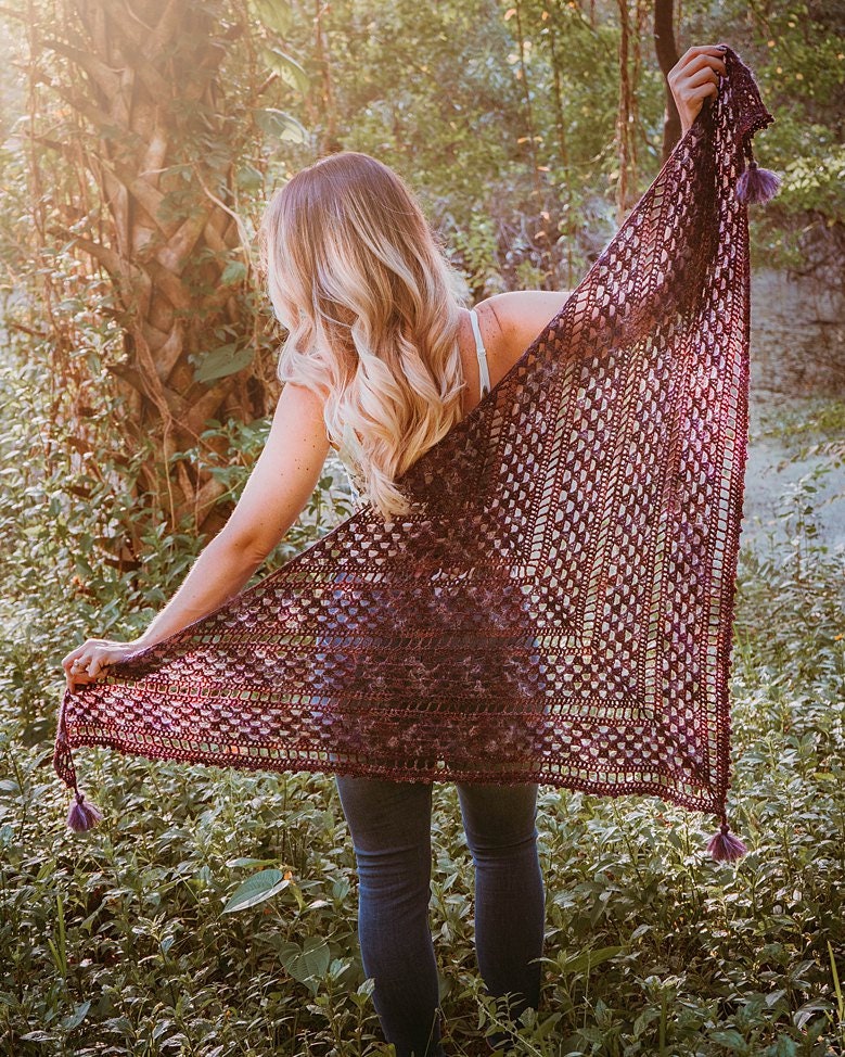 Amidst the lush greenery, a person stands holding a large triangular crochet shawl with tassels, reminiscent of vintage clothes. Sunlight filters through the trees, casting enchanting patterns around them.
