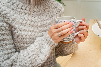 Close-up of a gray crochet sweater with braided puff stitches and hands holding a patterned mug