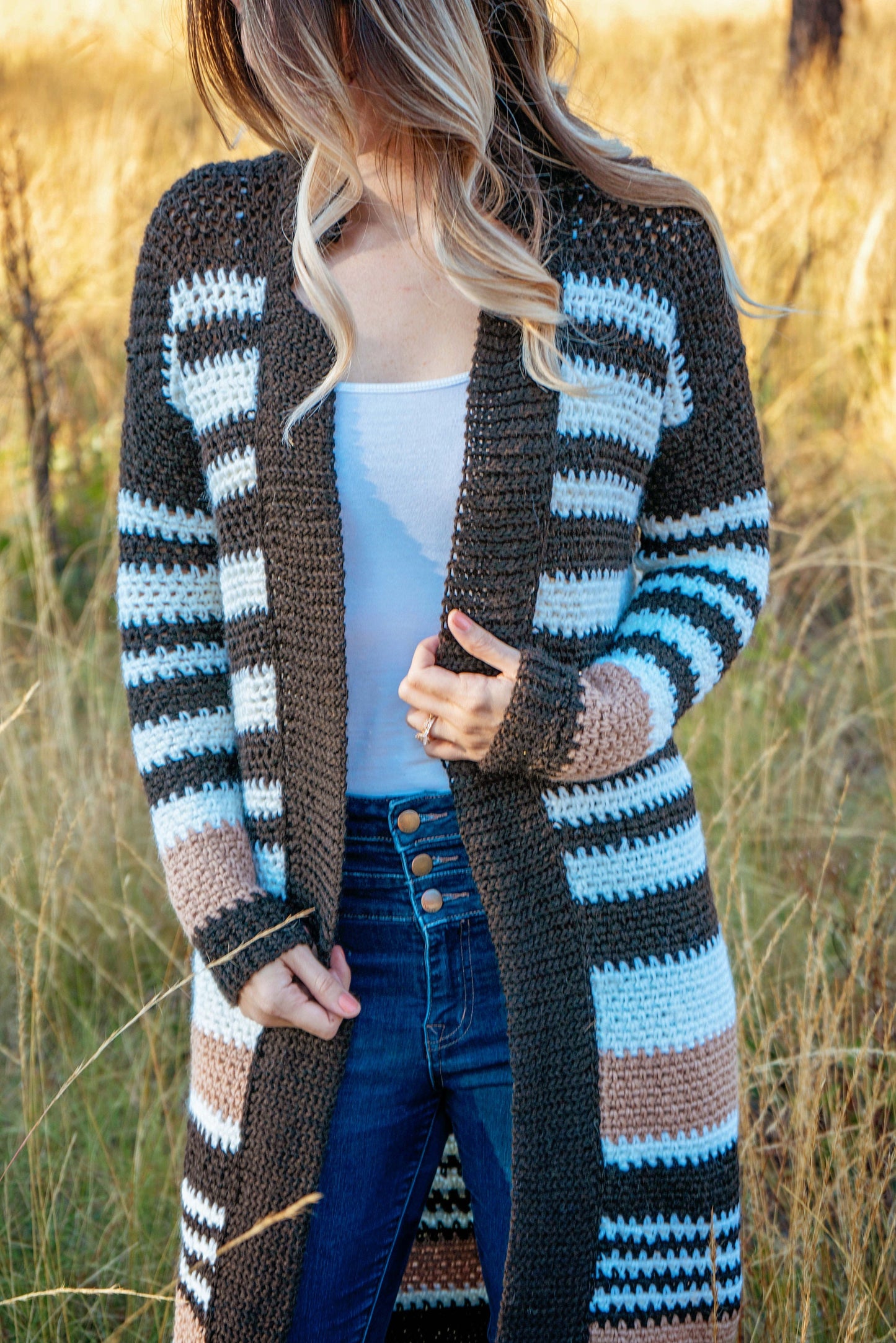 A person wearing a striped knit cardigan and blue jeans stands in a grassy field, adorned with unique jewellery that adds an extra touch of elegance to their vintage clothes.