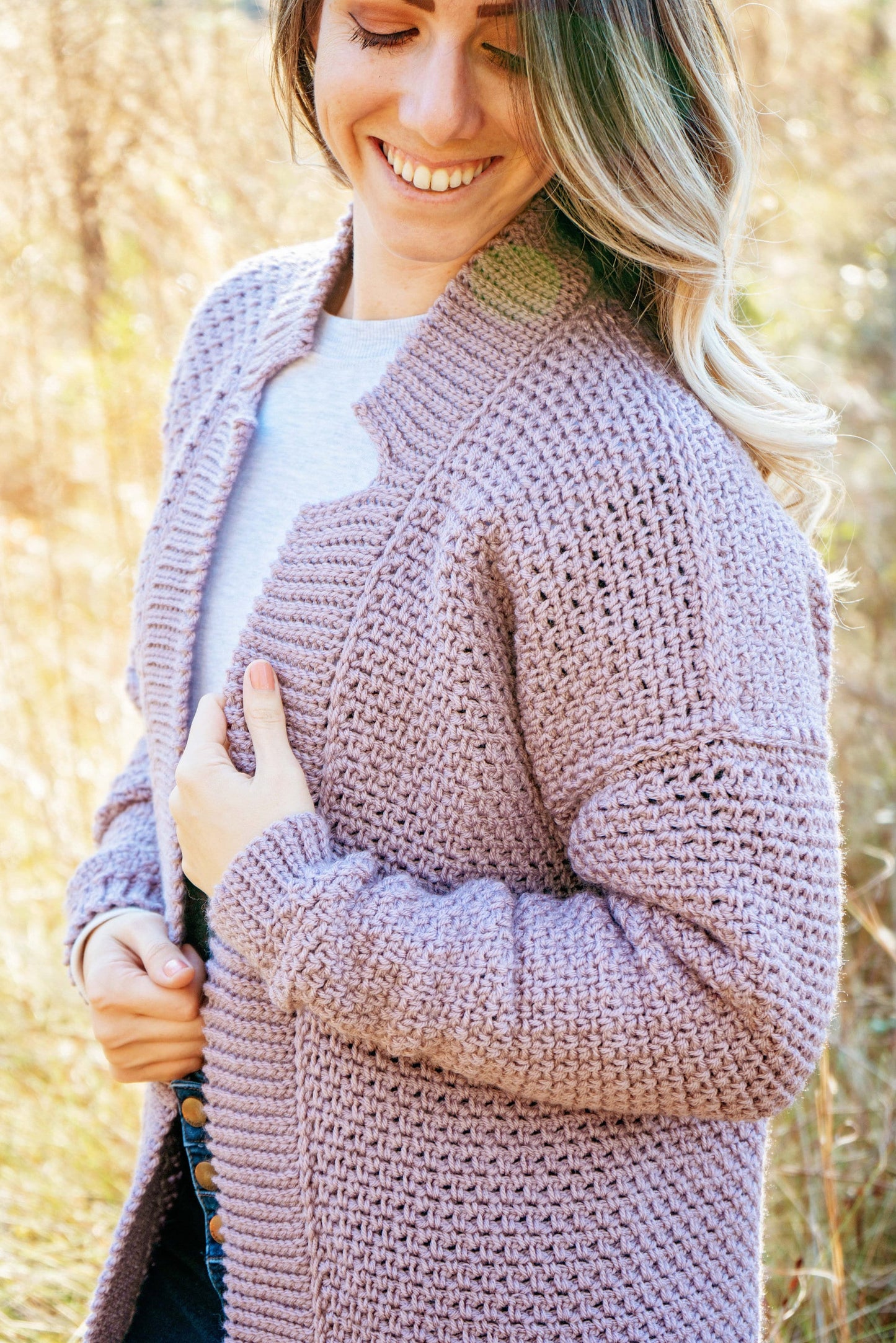 A person standing outdoors is wearing a light purple knitted cardigan over a white shirt and smiling while looking down. They are gently holding the cardigan with one hand, showcasing their vintage clothes with a touch of unique jewellery.